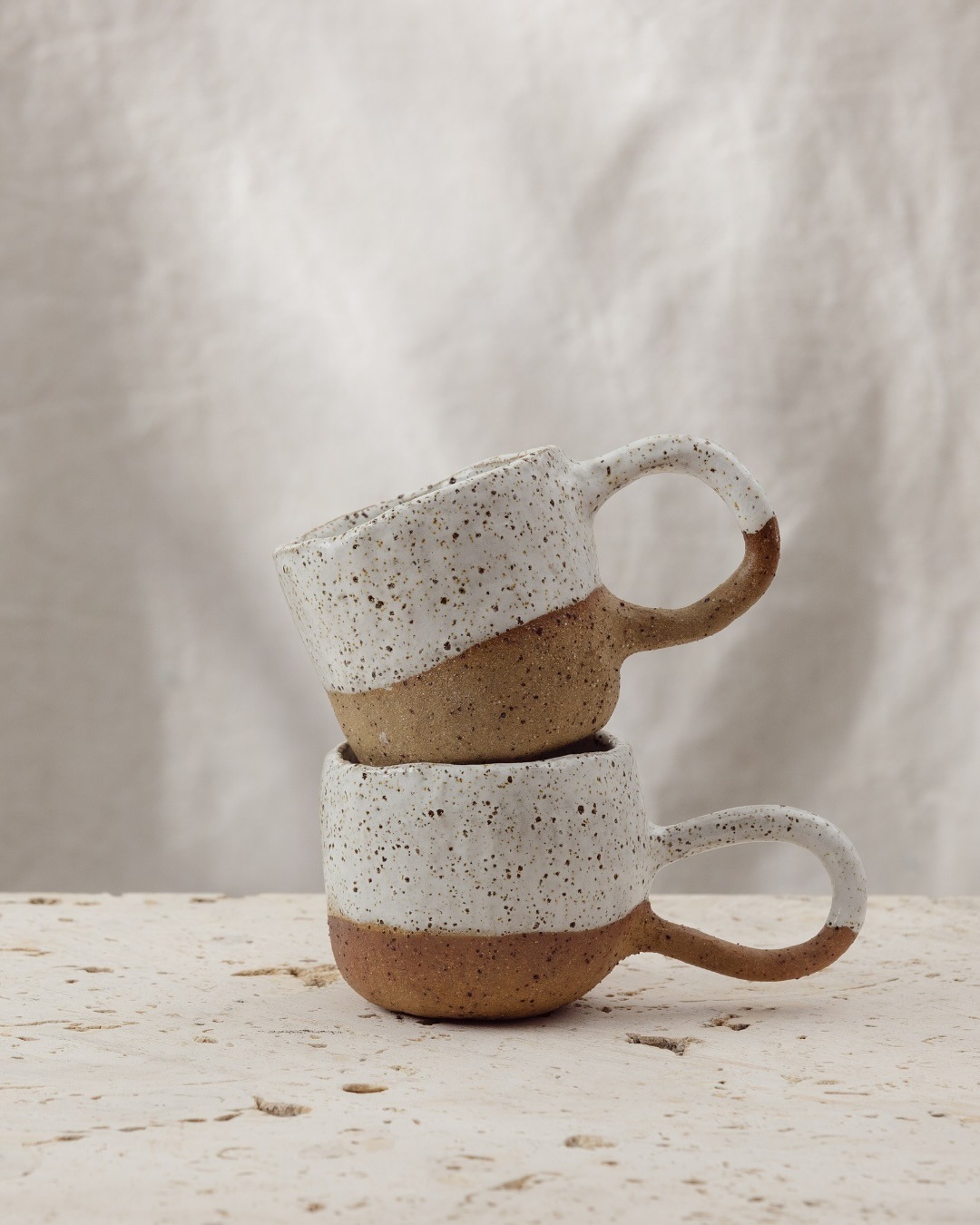 Two speckled mugs stacked on a travertine surface